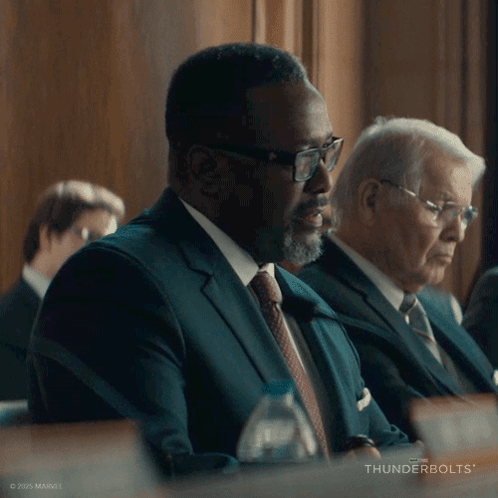 A Man In A Suit And Tie Is Sitting At A Table With Thunderbolts Written On The Bottom Right GIF