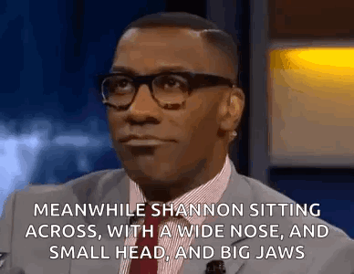 A Man In A Suit And Tie Is Sitting Down With A Wide Nose And A Small Head And Big Jaws . GIF