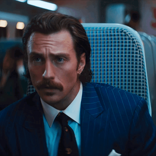 A Man In A Suit And Tie Is Sitting In A Blue Chair GIF