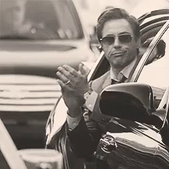 A Man In A Suit And Tie Is Sitting In A Car Waving His Hand . GIF