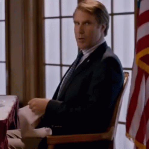 A Man In A Suit And Tie Is Sitting In A Chair Holding A Piece Of Paper . GIF