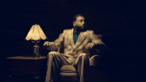 A Man In A Suit And Tie Is Sitting In A Chair In Front Of A Lamp GIF