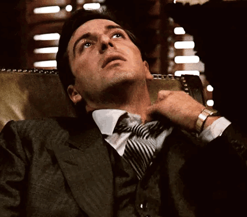 A Man In A Suit And Tie Is Sitting In A Chair GIF