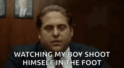 A Man In A Suit And Tie Is Sitting In A Chair Watching His Boy Shoot Himself In The Foot . GIF