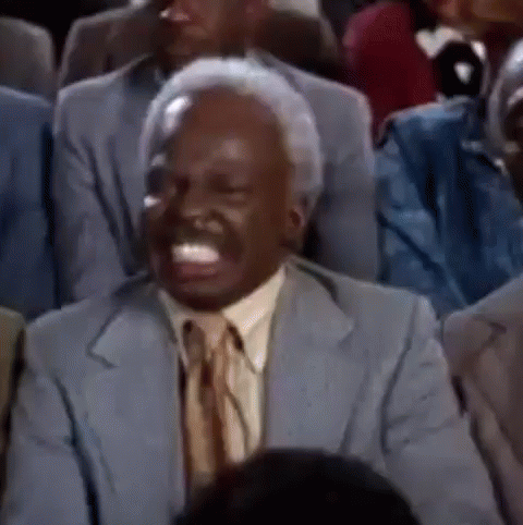 A Man In A Suit And Tie Is Sitting In A Crowd Of People And Laughing . GIF