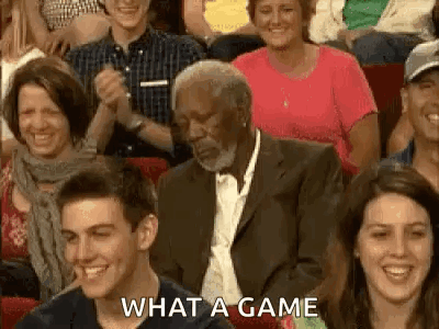 A Man In A Suit And Tie Is Sitting In A Crowd Of People Laughing . GIF