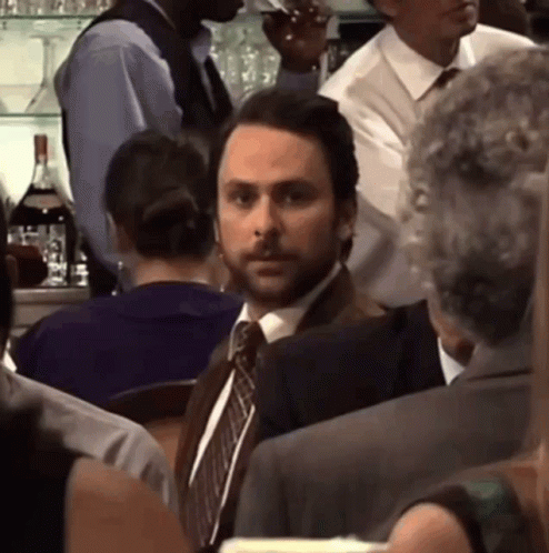 A Man In A Suit And Tie Is Sitting In A Crowded Room GIF