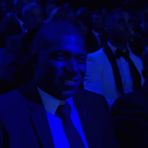 A Man In A Suit And Tie Is Sitting In A Dark Room Under Blue Lights GIF