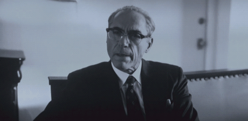 A Man In A Suit And Tie Is Sitting In A Room GIF