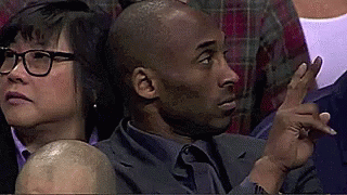 A Man In A Suit And Tie Is Sitting In A Stadium With A Woman Behind Him . GIF