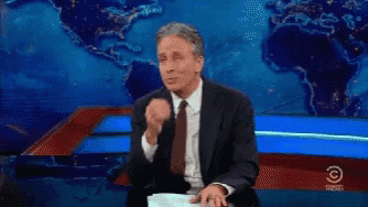 A Man In A Suit And Tie Is Sitting In Front Of A Comedy Central Sign GIF