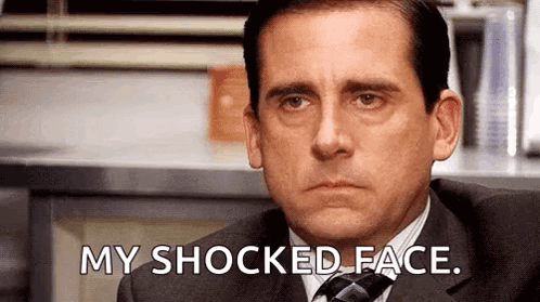 A Man In A Suit And Tie Is Sitting In Front Of A Counter And Says `` My Shocked Face '' . GIF