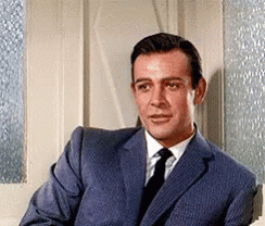 A Man In A Suit And Tie Is Sitting In Front Of A Door . GIF