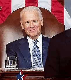 A Man In A Suit And Tie Is Sitting In Front Of A Flag . GIF