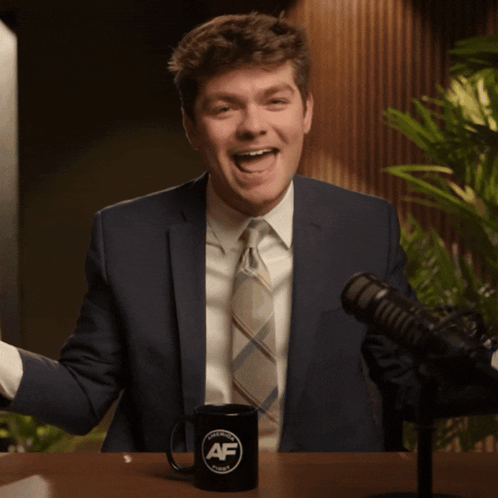 A Man In A Suit And Tie Is Sitting In Front Of A Microphone And A Mug That Says Af GIF