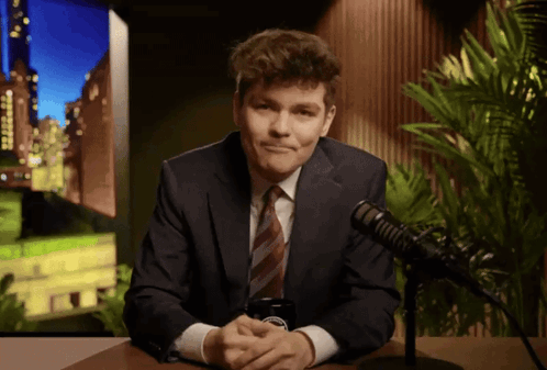 A Man In A Suit And Tie Is Sitting In Front Of A Microphone With A Cell Phone In His Hand GIF