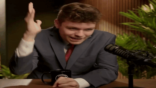 A Man In A Suit And Tie Is Sitting In Front Of A Microphone With A Coffee Mug On The Table GIF