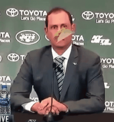 A Man In A Suit And Tie Is Sitting In Front Of A Microphone With A Toyota Logo Behind Him GIF