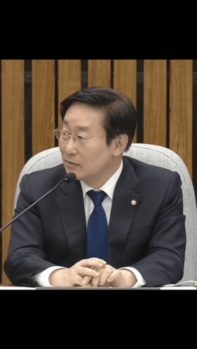 A Man In A Suit And Tie Is Sitting In Front Of A Microphone With His Hands Folded GIF