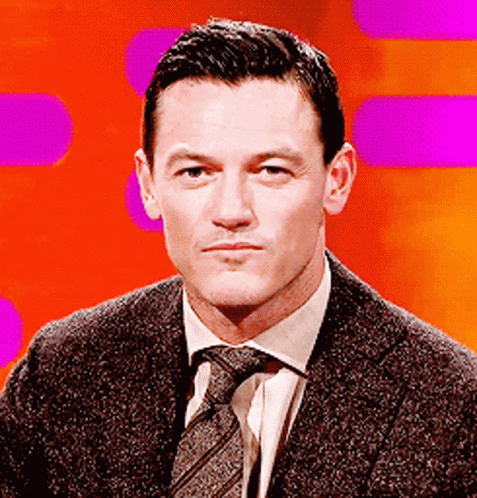 A Man In A Suit And Tie Is Sitting In Front Of A Purple Background GIF