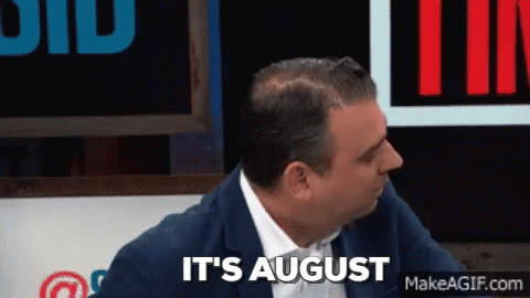 A Man In A Suit And Tie Is Sitting In Front Of A Sign That Says It 'S August GIF