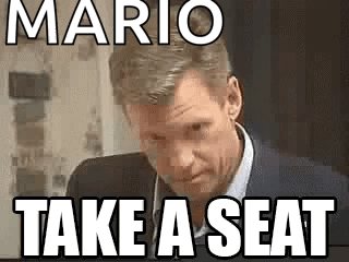 A Man In A Suit And Tie Is Sitting In Front Of A Sign That Says Mario Take A Seat . GIF