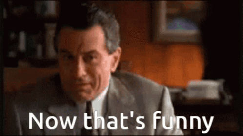 A Man In A Suit And Tie Is Sitting In Front Of A Sign That Says Now That 'S Funny GIF