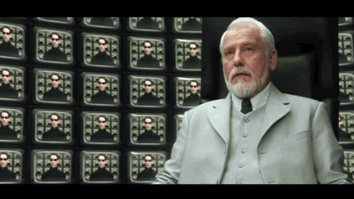 A Man In A Suit And Tie Is Sitting In Front Of A Wall Of Television Screens GIF