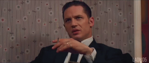 A Man In A Suit And Tie Is Sitting In Front Of A Wall With A Floral Pattern GIF