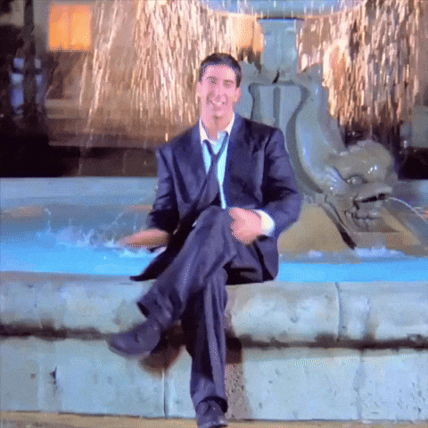 A Man In A Suit And Tie Is Sitting In Front Of A Water Fountain GIF