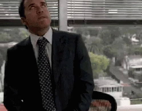 A Man In A Suit And Tie Is Sitting In Front Of A Window And Looking Up . GIF