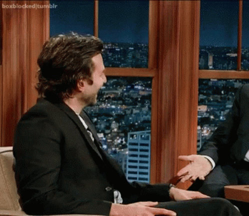 A Man In A Suit And Tie Is Sitting In Front Of A Window Talking To Another Man .. GIF