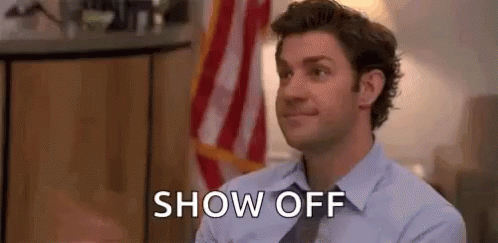 A Man In A Suit And Tie Is Sitting In Front Of An American Flag And Says `` Show Off '' . GIF