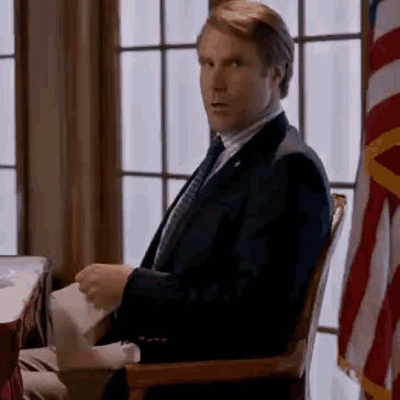 A Man In A Suit And Tie Is Sitting In Front Of An American Flag . GIF
