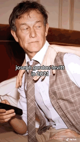A Man In A Suit And Tie Is Sitting On A Bed With A Woman Holding His Arm . GIF