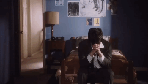 A Man In A Suit And Tie Is Sitting On A Bed With His Hands Folded . GIF
