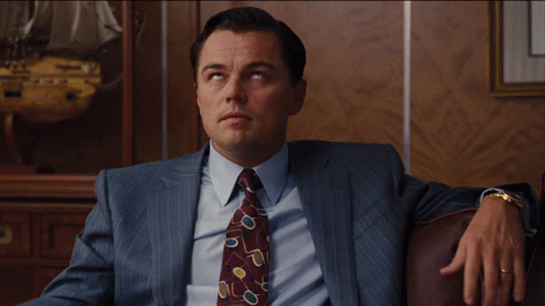 A Man In A Suit And Tie Is Sitting On A Brown Couch GIF