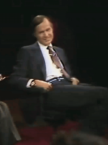 A Man In A Suit And Tie Is Sitting On A Chair GIF