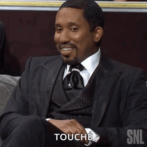 A Man In A Suit And Tie Is Sitting On A Couch And Touching His Chest With The Snl Logo Behind Him GIF