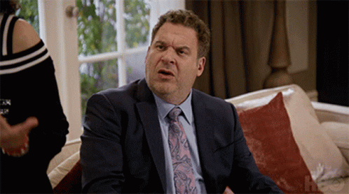 A Man In A Suit And Tie Is Sitting On A Couch With A Woman Behind Him GIF