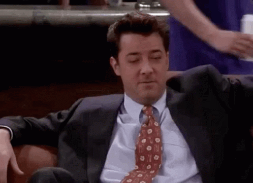A Man In A Suit And Tie Is Sitting On A Couch With His Hands On His Hips . GIF