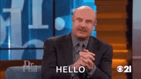 A Man In A Suit And Tie Is Sitting On A Stage With His Hands Folded And Saying Hello . GIF