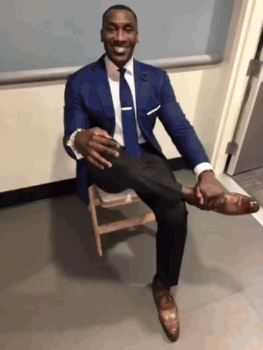 A Man In A Suit And Tie Is Sitting On A Wooden Chair With His Leg Up GIF