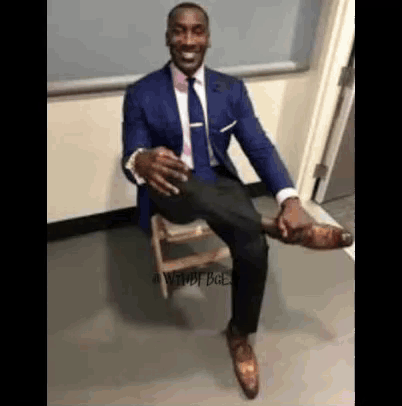 A Man In A Suit And Tie Is Sitting On A Wooden Stool With His Leg Up . GIF