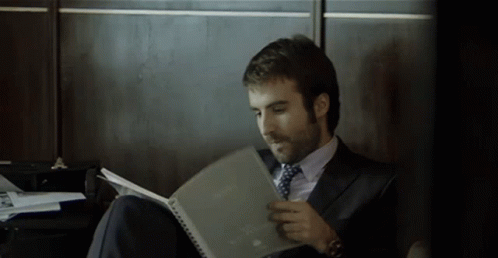 A Man In A Suit And Tie Is Sitting On The Floor Reading A Book GIF