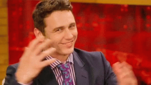 A Man In A Suit And Tie Is Smiling And Clapping His Hands In Front Of A Red Curtain . GIF
