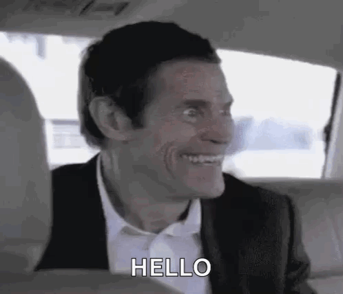 A Man In A Suit And Tie Is Smiling And Saying Hello While Sitting In The Back Seat Of A Car . GIF