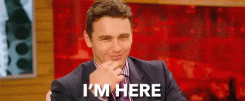 A Man In A Suit And Tie Is Smiling And Saying `` I 'M Here '' While Sitting On A Stage . GIF