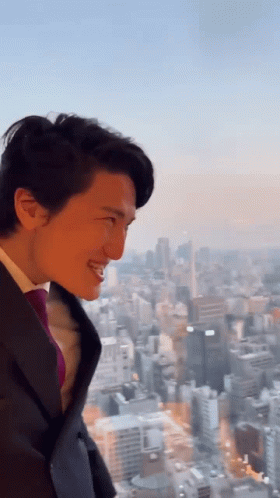 A Man In A Suit And Tie Is Smiling In Front Of A City Skyline GIF