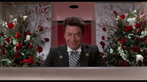 A Man In A Suit And Tie Is Smiling In Front Of A Display Of Flowers GIF